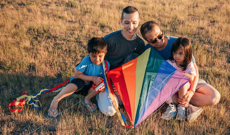 Two dads holding rainbow kite with their young children in a field