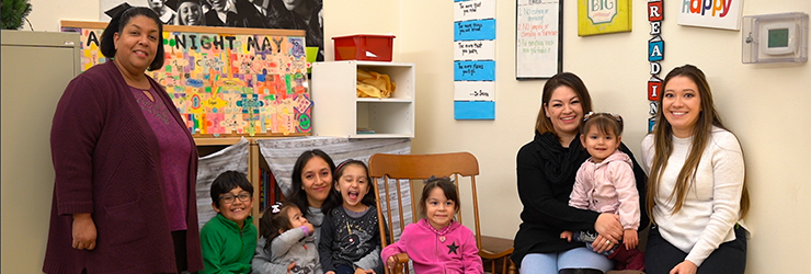 Two mothers and their five children, along with two female staff members, gathering and smiling in the community room at Mercy House Northwest Appian Way apartments