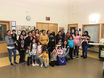 12 adults and 16 children of varying ages are standing together in a large, brightly lit community room inside the Mercy Housing Appian Way building.