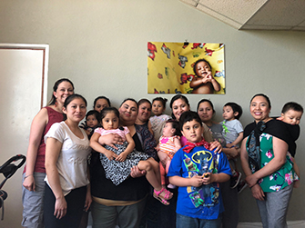 Six mothers and their six children are standing in a group, under a photo of a smiling baby, all facing the camera. On the right side is the Open Arms Perinatal Services doula, who served as a co-facilitator carrying her son on a wrap-style toddler carrier sling. On the left stands the PEPS East Region Community Connector, who also served as a co-facilitator for this group of women. 